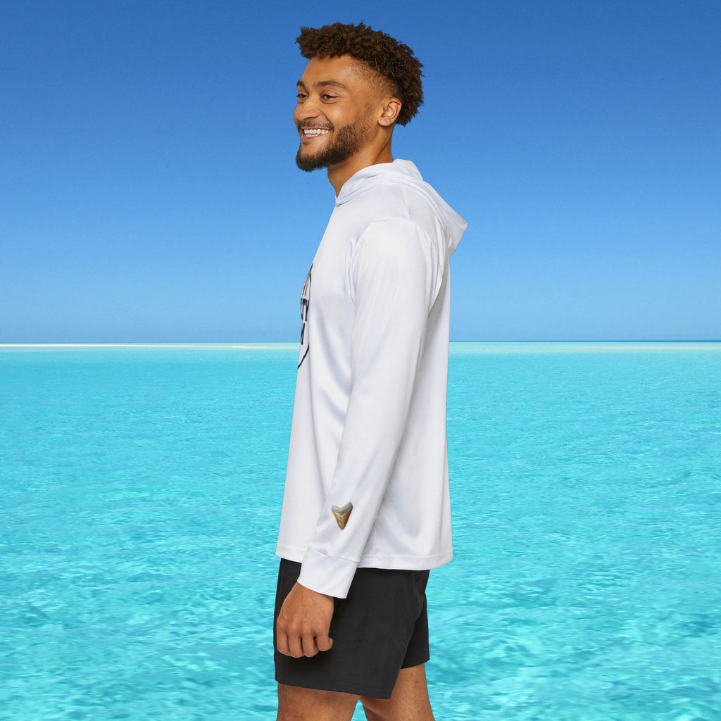 Man wearing a white long-sleeve shirt and black shorts standing on a beach with clear blue water and sky.