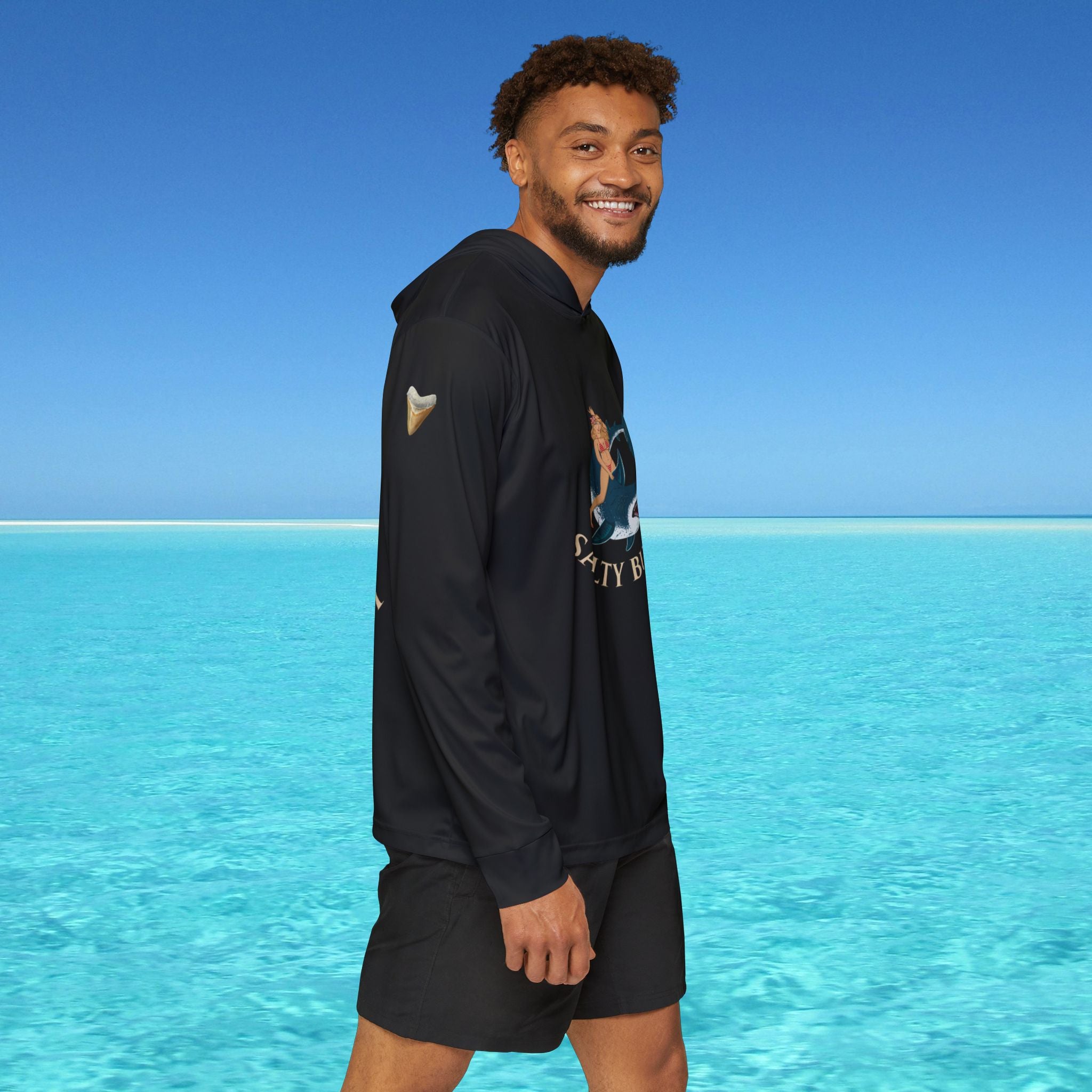 Man wearing a black hoodie and shorts standing in front of clear blue water and sky.
