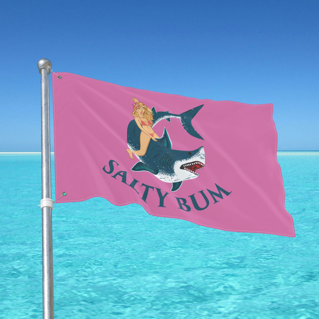 Pink flag with a graphic of a woman on a shark and 'Salty Bum' text, waving in front of a blue sky and ocean.