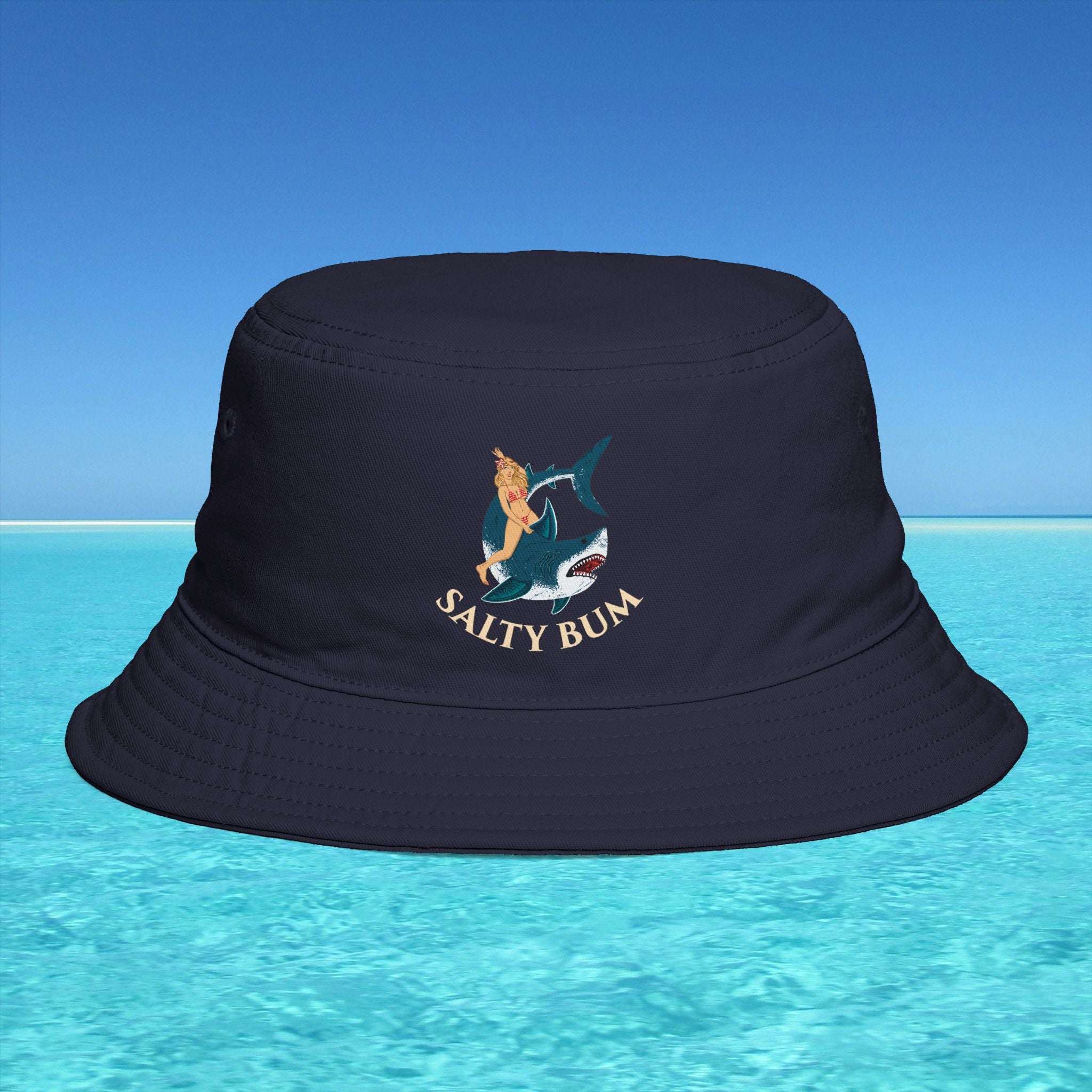 Navy bucket hat with 'Salty Bum' logo on a blue water and sky background