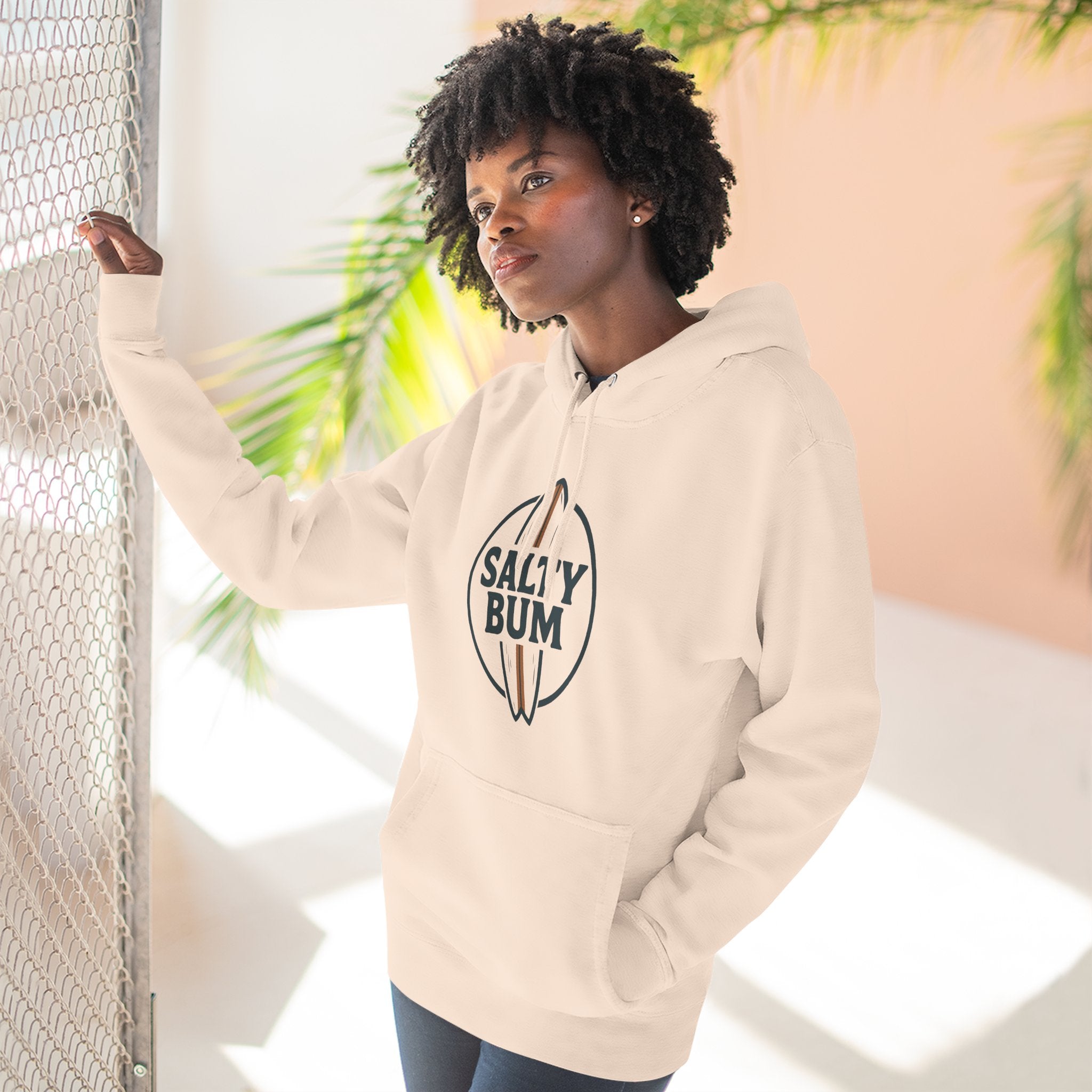 Woman wearing a beige hoodie with 'Salty Bum' logo, standing against a light background with plants.