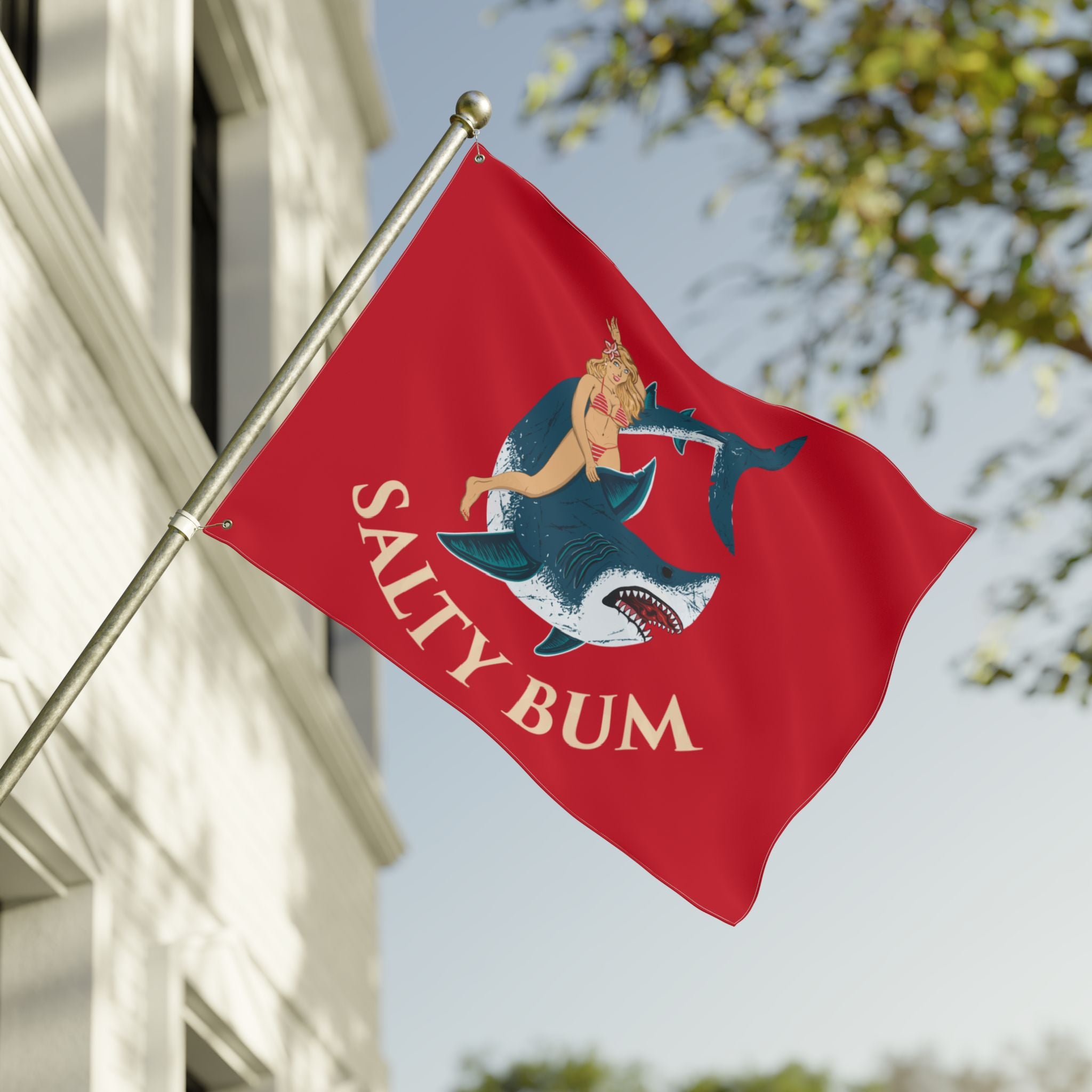 Red flag with a shark and woman design and 'Salty Bum' text, waving in front of a building.