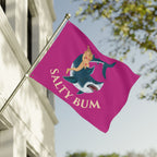 Flag with a shark and bikini-clad figure design on a pink background, featuring the text 'Salty Bum', waving in front of a building.