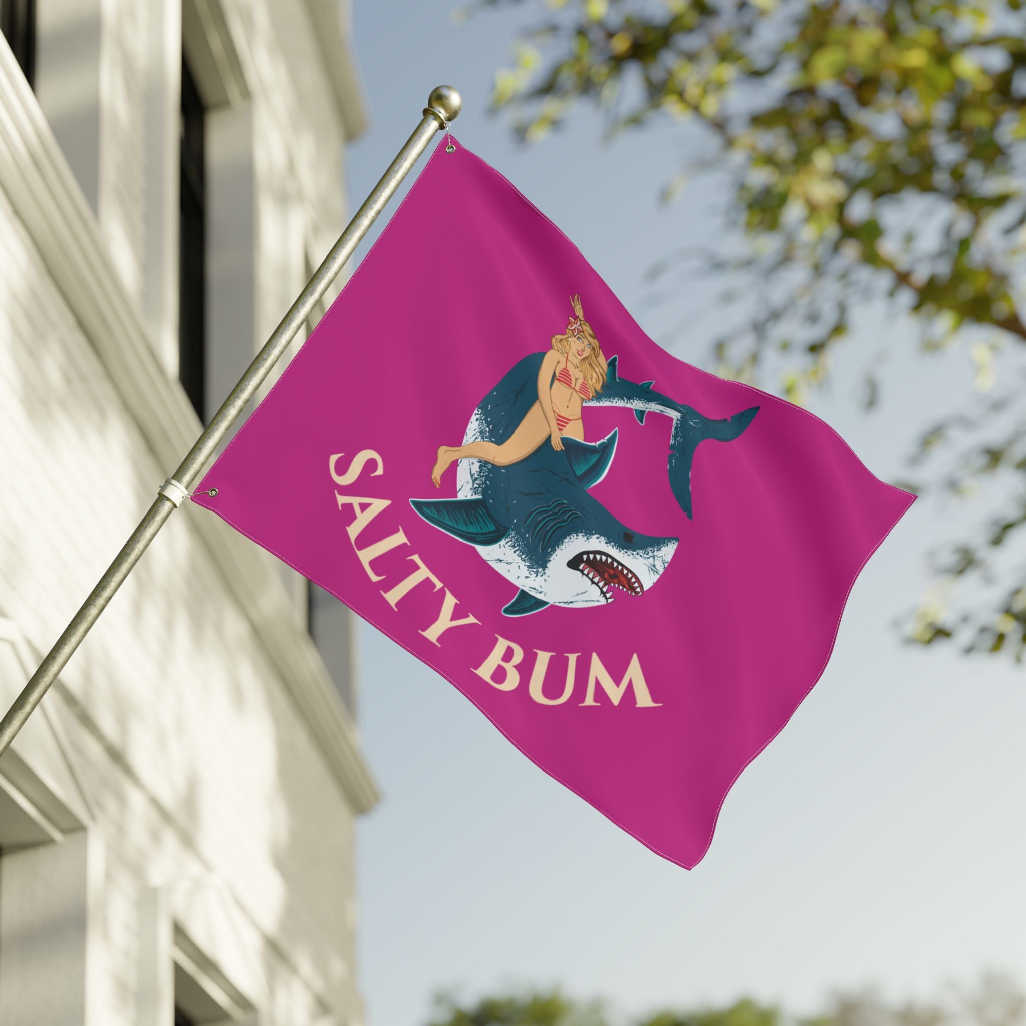 Flag with a shark and bikini-clad figure design on a pink background, featuring the text 'Salty Bum', waving in front of a building.