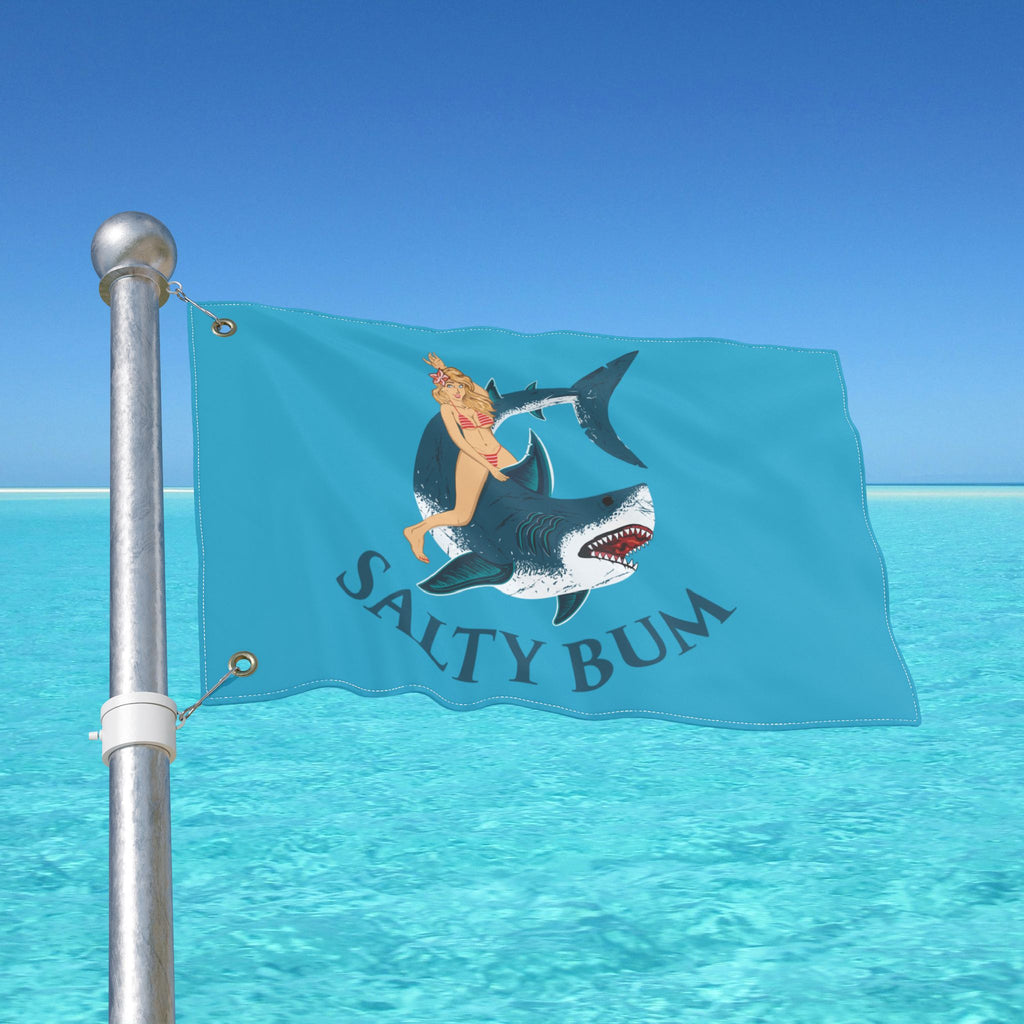 Flag with a graphic of a person on a shark and 'Salty Bum' text, against a clear blue sky.