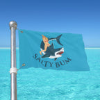 Flag with a graphic of a person on a shark and 'Salty Bum' text, against a clear blue sky.