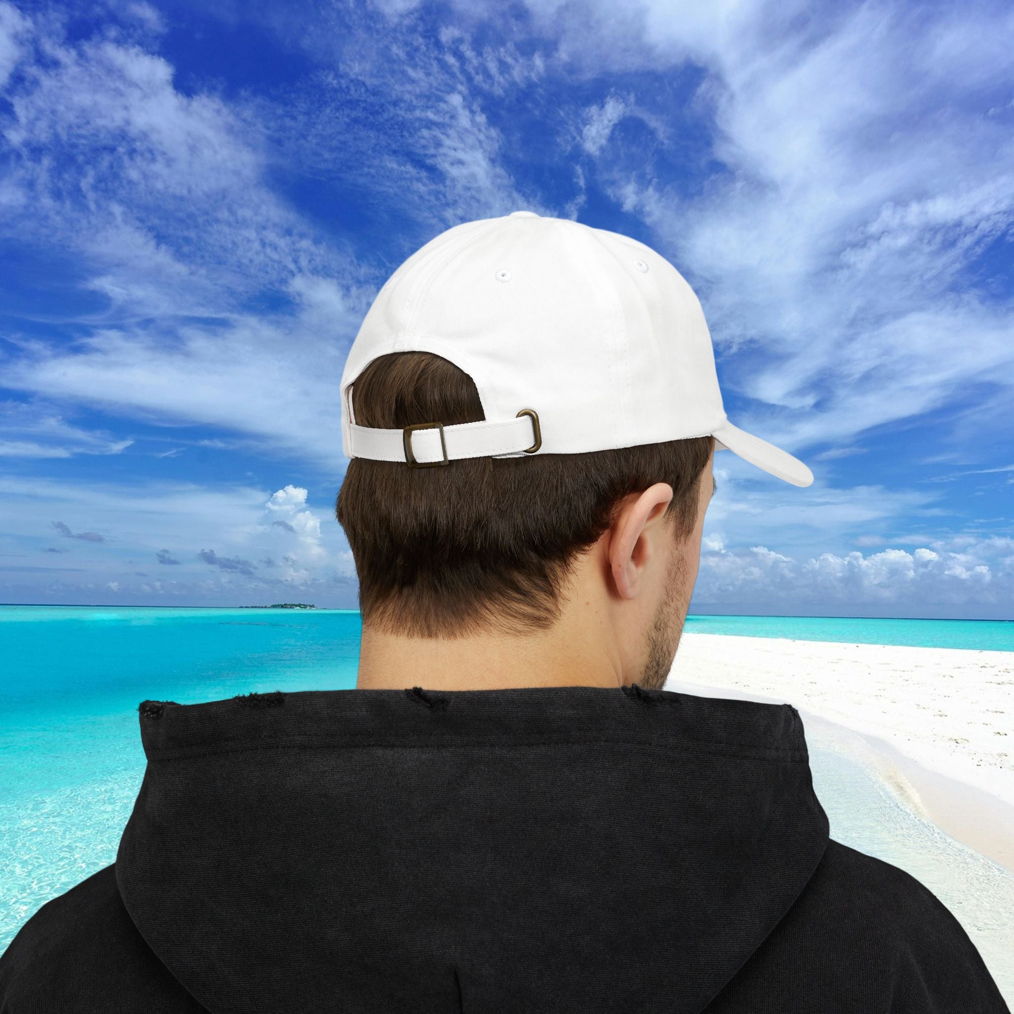 Person wearing a white cap and black hoodie looking at a tropical beach with clear blue water and sky.