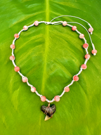A beaded necklace with round pinkish-orange beads on a white cord, featuring a dark shark tooth pendant wrapped with copper wire, displayed on a large green leaf.