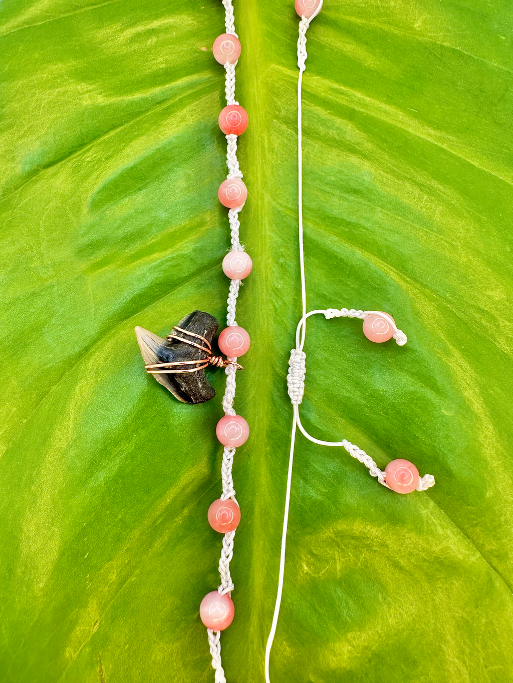 A handmade beaded necklace and matching earrings laid out on a large green leaf. The beads are a soft pinkish-orange, strung on white cord, with one dark stone and a small wrapped crystal accent.