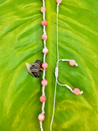 A handmade beaded necklace and matching earrings laid out on a large green leaf. The beads are a soft pinkish-orange, strung on white cord, with one dark stone and a small wrapped crystal accent.